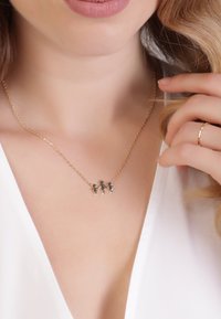 Halskette in Golfarbe mit drei silbernen Figuren, die miteinander verbunden sind. Die Kette ist zart und ergänzt ein V-Ausschnitt-Oberteil.
