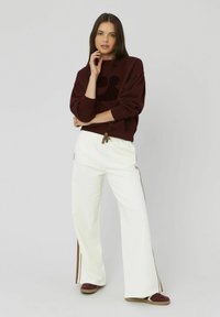 Sweatshirt bordeaux avec un grand graphique noir sur le devant, pantalon large blanc avec des rayures latérales marron et des chaussures bordeaux foncé.