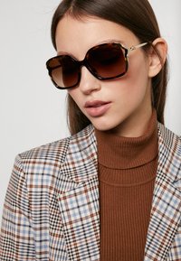 Occhiali da sole tartarugati con montatura oversized e tondeggiante; lenti marroni sfumate; dettagli dorati sulle tempie. Indossati con un dolcevita marrone a coste e un blazer a quadri.