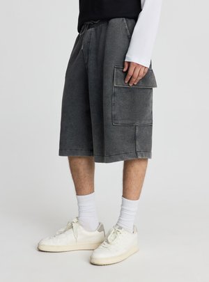 Persona che indossa pantaloncini cargo grigio scuro fino al ginocchio, calzini bianchi e sneakers bianche, in piedi davanti a uno sfondo chiaro uniforme.
