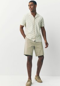 Kortærmet lysegrøn skjorte med blomster mønster, parret med beige shorts og brune sko. Modellen står med hænderne i lommerne.