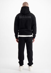 Zwarte hoodie met een zak aan de voorkant, groot logo op de achterkant, gecombineerd met zwarte sweatpants en witte sneakers. Gemaakt van een katoenmix.