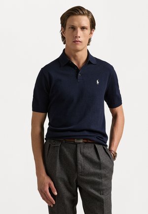 Navy polo shirt in cotton with a three-button placket and small white embroidered logo; paired with gray wool trousers featuring pleats.