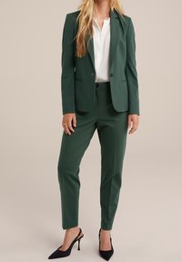 Groene op maat gemaakte blazer en bijpassende broek van gladde stof, gecombineerd met een witte blouse. Zwarte hoge hakken maken de outfit compleet.