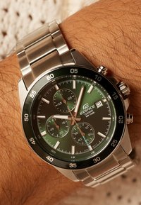 Zilverkleurig horloge van roestvrij staal met een groene gradient wijzerplaat, chronograaf subwijzerplaten en zwarte lunette. Voorzien van lichtgevende uurmarkeringen en datumweergave.