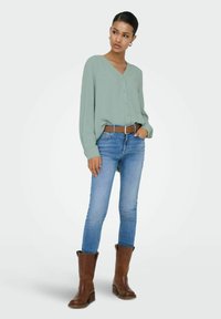 Femme portant un chemisier vert sauge boutonné, un jean skinny bleu avec une ceinture marron, et des bottes en cuir marron mi-mollet, se tenant devant un fond blanc.