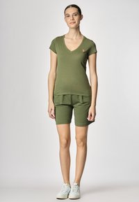 T-shirt in cotone verde oliva a V con maniche corte abbinato a pantaloni corti coordinati. Dettaglio della tasca sul pantalone corto in evidenza. Indossato con sneaker bianche.