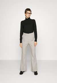 Un pull à col roulé noir, un pantalon flare taille haute gris clair et des chaussures noires. Le mannequin porte de grandes lunettes rectangulaires et se tient droit.