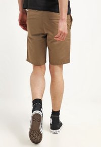 Pantalones cortos caqui de corte recto, con dos bolsillos traseros y una textura suave, combinados con zapatillas de deporte negras de caña alta y calcetines hasta el tobillo.