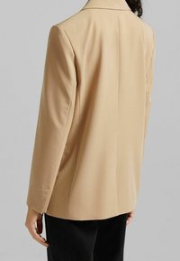 Beżowa marynarka dopasowana z klapami, pojedynczym rozcięciem z tyłu i gładkiego materiału; w zestawieniu z czarnymi spodniami dla kontrastu.