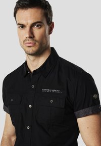 Camisa negra de manga corta con cierre de botones, dos bolsillos en el pecho y detalles en tela gris en los puños. Cuenta con bordado de la marca y botones a presión elegantes.