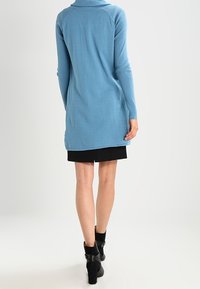 Světle modrý kardigan s dlouhým rukávem a vysokým límcem, s vroubkovanou texturou a mírně zakřiveným lemem, kombinovaný s černou sukní a kotníkovými botami.