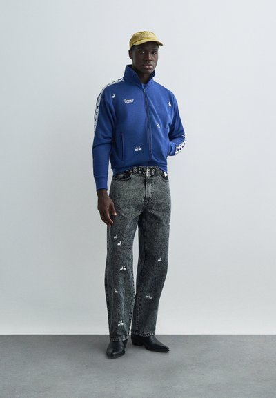 Zils zip-up džemperis ar baltām etiķetēm un sānu svītrām, kopā ar nodilušām melnām džinsām ar baltām izšūtām detaļām. Melni zābaki.