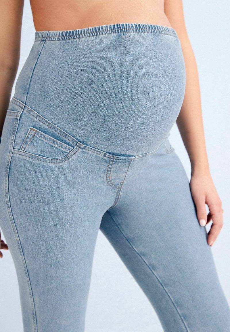 Jeans de maternidad azul claro con cintura elástica, diseño ajustado y bolsillos frontales diagonales. Textura de denim suave, silueta ajustada.