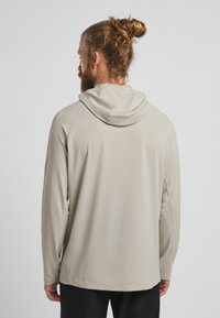 Haut à capuche beige clair à manches longues avec un cordon de serrage, texture lisse et coutures à plat. Le dos présente un léger design à ourlet droit.