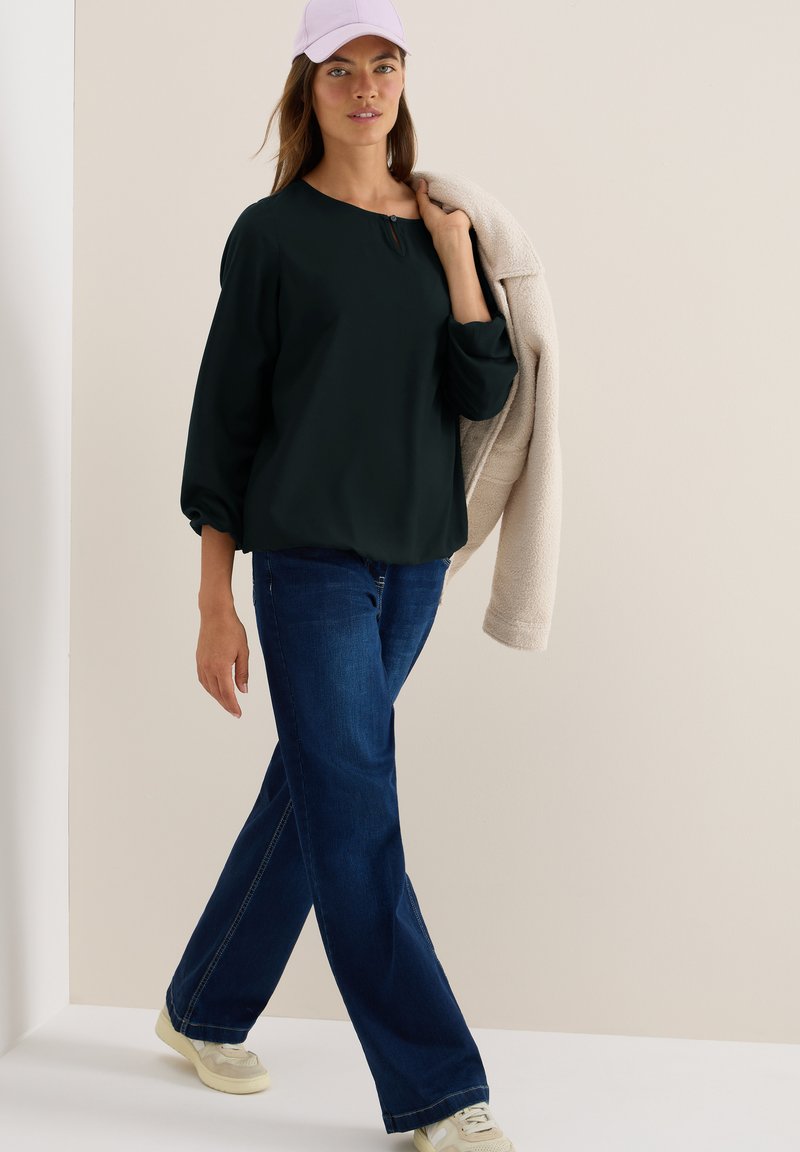 Vrouw die loopt in een zwart blouse, donkerblauwe jeans, witte sneakers, een lavendelcap en een beige teddyjas over haar schouder draagt.