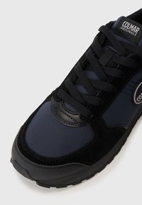 Black and navy athletic shoe featuring a combination of smooth synthetic, velvet-like texture, round toe, padded tongue, and logo accent.