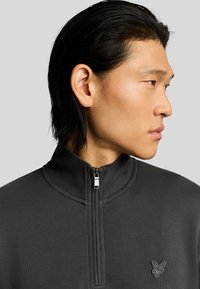 Felpa zip con collo alto di colore grigio scuro, caratterizzata da una texture di tessuto liscia e un sottile logo del marchio sul petto. Dettaglio della zip in argento.