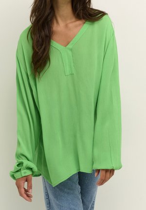 Woman wearing loose-fitting, long-sleeve green blouse with V-neck and light blue jeans, standing against a plain light background.