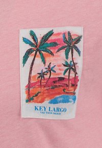 Różowa koszulka z prostokątną naszywką przedstawiającą tropikalny krajobraz z palmami, zachodem słońca w tle oraz napisem "NASTRÓJ WAKACYJNY KEY LARGO."