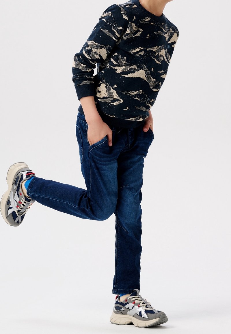 Enfant portant un pull à motif sombre, un jean bleu et des baskets, avec une jambe pliée en arrière, les mains dans les poches, debout sur un fond blanc.