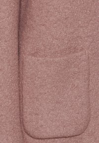 Rosa texturerat yttertyg med en synlig påsydd ficka. Ytan verkar mjuk och ludden, med en enhetlig färg genom hela tygstycket.