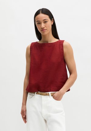 Kobieta z długimi ciemnymi włosami, ubrana w czerwoną bezrękawnik i białe spodnie z jasnobrązowym paskiem, stoi z jedną ręką w kieszeni.