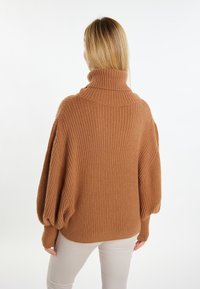 Pulover supradimensionat, cu guler înalt cu nervuri, de culoare maro închis. Prezintă mâneci lejere și tricotaj texturat. Asortat cu pantaloni de culoare deschisă.