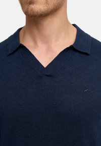 Marineblauer Pullover mit V-Ausschnitt und kurzem Kragen, aus einem glatten Strickmaterial gefertigt. Verfügt über ein dezentes gesticktes Logo auf der Brust.