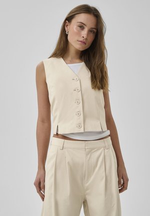 Ung kvinde med langt brunt hår iført cremefarvet, knappet, ærmeløs vest og matchende bukser med højt talje, stående foran en grå baggrund.