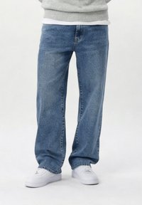 Άτομο που φορά χαλαρά μπλε τζιν και λευκά αθλητικά παπούτσια, στέκεται μπροστά σε απλό φωτεινό φόντο.