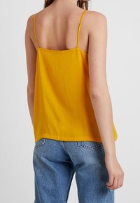Top amarillo sin mangas con tirantes finos, hecho de una tela suave. Corte holgado, dobladillo recto, diseño minimalista. Combinado con jeans de color azul claro.