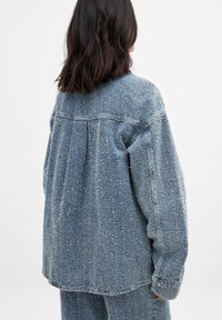 Oversized-Denimjacke in Hellblau mit strukturierter Oberfläche, mit gesenkten Schultern und einem Rückenschlitz für zusätzliche Bewegungsfreiheit.
