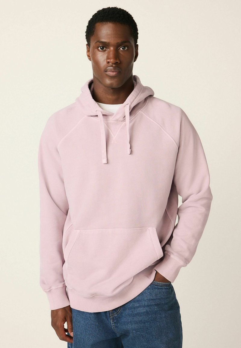 Jeune homme portant un sweat à capuche rose clair et un jean bleu, debout avec une main dans la poche devant un fond uni clair.