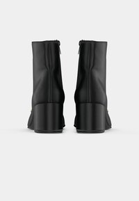 Bottines noires en matériau synthétique lisse avec un bout arrondi, un haut talon carré et une fermeture éclair visible à l'arrière.