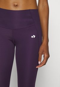 Fialové legíny s širokým pasem, hladkou texturou a jemným bílým logem na levé straně. Detailní záběr na pas a ruku.