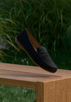 Mocassin en daim noir avec un bout arrondi, des détails cousus et un accent en relief sur le dessus. Présenté sur un fond vert naturel.