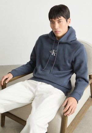 Sweat à capuche bleu marine avec cordons, arborant un logo sur la poitrine, associé à un pantalon blanc, assis sur une chaise en bois.