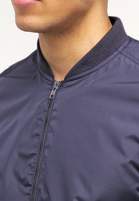 Giacca bomber blu navy realizzata in un materiale liscio e lucido, con colletto a costine e zip frontale, dal taglio affusolato.