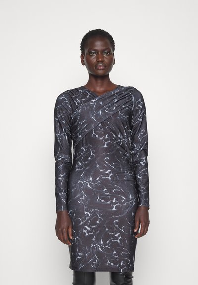 Žena v dlhých rukávoch, v čiernych šatách po kolená s bielym mramorovým vzorom, stojí pred jednoduchým bielym pozadím.