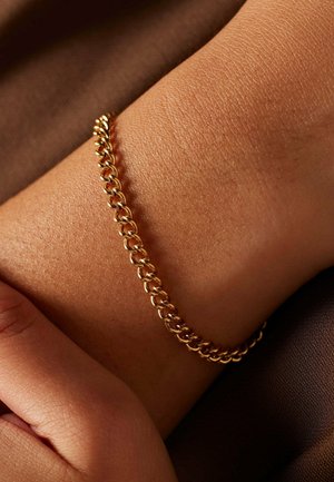 Gold chain bracelet on a wrist, featuring interlocking links with a smooth finish. Background shows a textured brown fabric.