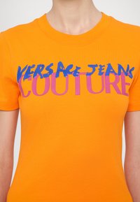 Oranžové bavlněné tričko s kulatým výstřihem. Obsahuje text "VERSACE JEANS COUTURE" v modré a růžové barvě s efektem štětcem.