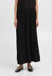 Jupe maxi noire avec une taille élastique, en tissu doux et à la coupe fluide, tombant jusqu'aux chevilles, assortie à des ballerines noires.