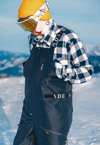 Personne portant un bonnet jaune, des lunettes de ski, une chemise à carreaux et une salopette de neige noire, debout sur une montagne enneigée avec un arrière-plan flou.