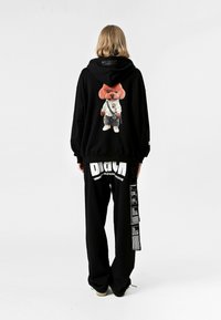 Sudadera negra de gran tamaño con un gráfico colorido de un oso vestido de artes marciales en la parte trasera. Combinada con pantalones negros y zapatillas.