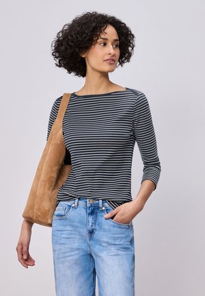 Frau mit lockigem Haar, die ein schwarz-weiß gestreiftes Langarmshirt, hellblaue Jeans trägt und eine beigefarbene Wildleder-Schultertasche bei sich hat.