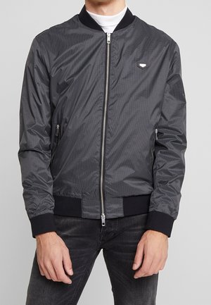 Mann trägt eine eng anliegende Bomberjacke in Schwarz und Grau kariert mit schwarzen Bündchen, Kragen und Reißverschluss über einem weißen Rollkragenpullover.