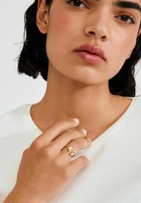 Kuldne sõrmus, millel on lillemotiivid, kantud sõrmel. Käsi toetub valgele soonikriidest taustale.