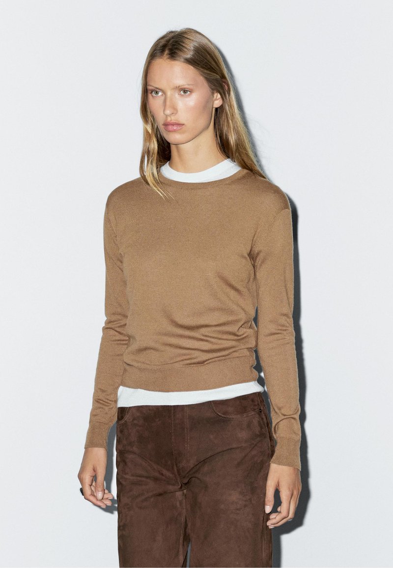 Brauner Strickpullover mit langen Ärmeln und rundem Ausschnitt, über einem weißen Hemd getragen. Weiche Textur, tailliert.
