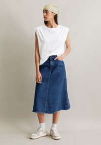 Witte mouwloze T-shirt, denim A-lijn rok en beige sneakers. De rok heeft zichtbare stiksels en is van middellange lengte tot de kuit.